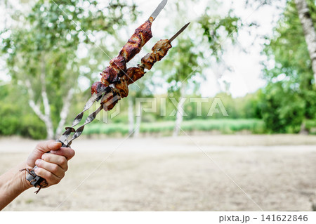 Grilled meat skewers held in hand outdoors. Delicious traditional barbecue shashlik on a blurry green background with wide copy space for text. Grilled meat skewers held in hand outdoors. Delicious traditional barbecue shashlik on a blurry green background with wide copy space for text. 141622846