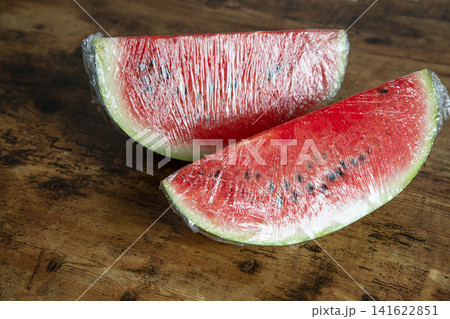 Two juicy red watermelon wedges covered with transparent stretch wrap on a rustic dark wooden table. Two juicy red watermelon wedges covered with transparent stretch wrap on a rustic dark wooden table. 141622851