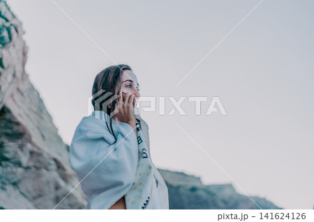 Woman, towel, coast. Young woman wrapped in a towel talks on phone at a rocky coast with copy space. 141624126