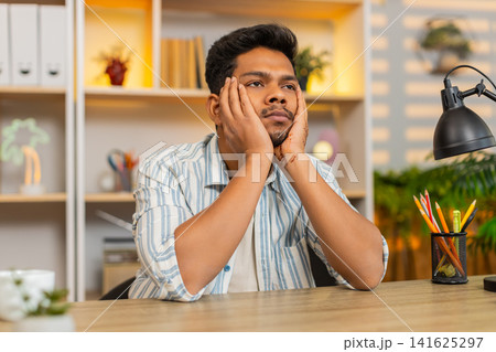 Indian man on home table leaning on hands looking exhausted after long remote work indoors at home 141625297