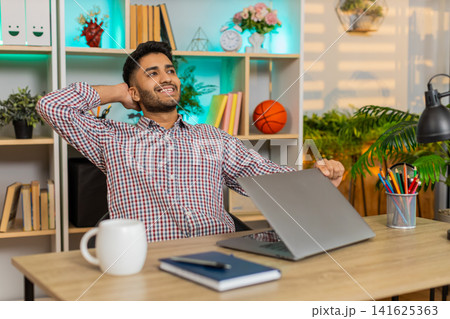 Indian man on home table closing laptop with satisfied smile after productive work routine indoors Indian man on home table closing laptop with satisfied smile after productive work routine indoors 141625363