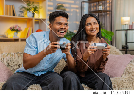 Indian couple man and woman sitting on home sofa play console games with wireless gamepads smiling Indian couple man and woman sitting on home sofa play console games with wireless gamepads smiling 141625391