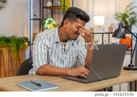 Indian man rubs eyes after long laptop work eye strain headache pain needs break and relief at desk Indian man rubs eyes after long laptop work eye strain headache pain needs break and relief at desk 141625556