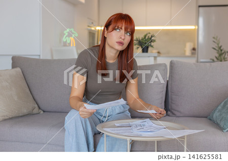 Young woman reviews paper bills and receipts frowns compares numbers checks figures verifies charges Young woman reviews paper bills and receipts frowns compares numbers checks figures verifies charges 141625581