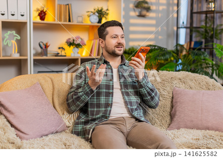 Young man on home sofa speaking on smartphone over loudspeaker with emotional hand gestures indoors 141625582