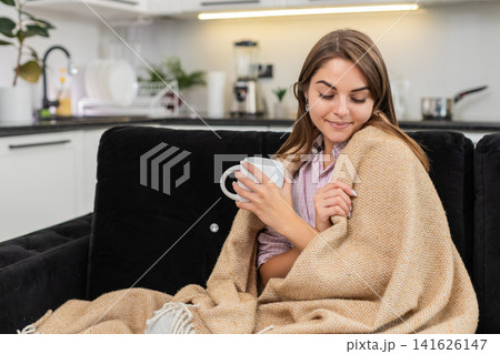 Young woman holds tea mug warms hands smiles under blanket enjoys quiet pause and peaceful deep rest Young woman holds tea mug warms hands smiles under blanket enjoys quiet pause and peaceful deep rest 141626147
