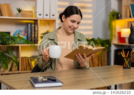 Young woman on home table reading book and drinking hot mug beverage during relaxing leisure indoors Young woman on home table reading book and drinking hot mug beverage during relaxing leisure indoors 141626210