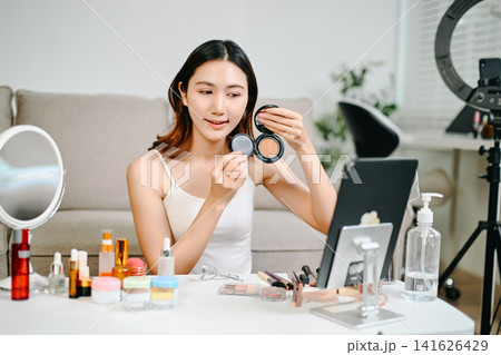 Asian beauty influencer filming a makeup tutorial using cosmetics, skincare products, smartphone and ring light in a modern home studio for social media content. Asian beauty influencer filming a makeup tutorial using cosmetics, skincare products, smartphone and ring light in a modern home studio for social media content. 141626429