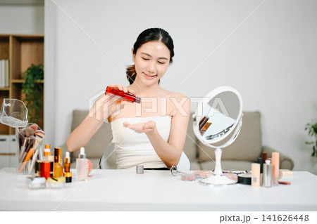 Asian woman practicing her daily skincare routine, gently touching her clear face while applying serum at a modern vanity. 141626448