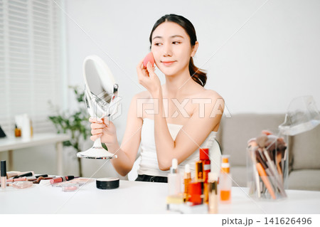 Young Asian woman applying makeup with eyeshadow palette while recording a beauty tutorial using smartphone and ring light in a modern home studio. Young Asian woman applying makeup with eyeshadow palette while recording a beauty tutorial using smartphone and ring light in a modern home studio. 141626496