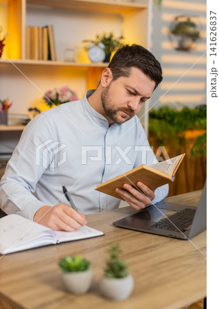 Young man on home table studying with laptop and book during online lesson and taking notes indoors Young man on home table studying with laptop and book during online lesson and taking notes indoors 141626837