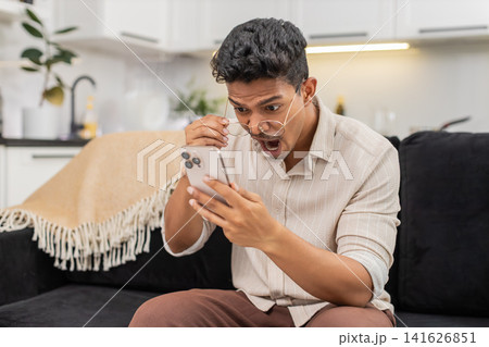 Indian man with glasses reads unexpected message on smartphone removes and puts glasses back laughs Indian man with glasses reads unexpected message on smartphone removes and puts glasses back laughs 141626851