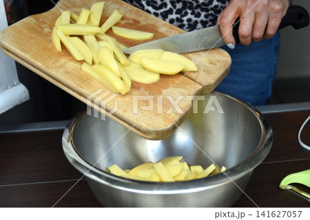 Close-up of a woman's hands transferring freshly cut homemade French fries from a wooden cutting board into a stainless steel bowl. Food preparation scene featuring fresh potatoes, home cooking. Close-up of a woman's hands transferring freshly cut homemade French fries from a wooden cutting board into a stainless steel bowl. Food preparation scene featuring fresh potatoes, home cooking. 141627057