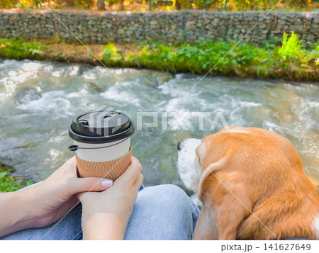 Unrecognizable woman hand holding a disposable coffee cup while sitting near her dog by a flowing river Unrecognizable woman hand holding a disposable coffee cup while sitting near her dog by a flowing river 141627649