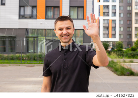 Man waving hand in greeting gesture. Casual portrait of a friendly guy smiling outdoors near modern residential buildings. Communication, welcome and social interaction concept. Man waving hand in greeting gesture. Casual portrait of a friendly guy smiling outdoors near modern residential buildings. Communication, welcome and social interaction concept. 141627822