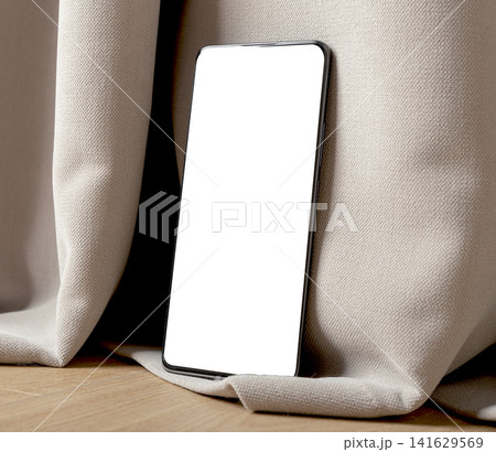 Blank smartphone mockup leaning against fabric on a wooden floor. Angled mobile phone with white screen in a clean indoor setting. Ideal for app design, advertising, and 141629569