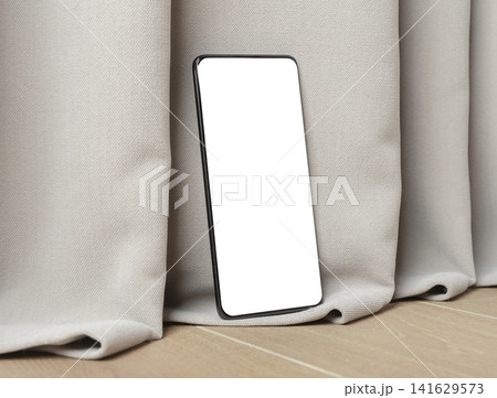 Blank smartphone mockup leaning on curtain fabric. Angled mobile phone with white screen in a minimalist interior. Ideal for app design, advertising, and product presentation.. 141629573