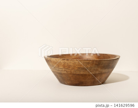 A rustic wooden bowl sits on a light beige surface photographed from a low side angle. The handcrafted bowl casts a soft shadow revealing its natural form and texture. Concept of A rustic wooden bowl sits on a light beige surface photographed from a low side angle. The handcrafted bowl casts a soft shadow revealing its natural form and texture. Concept of 141629596