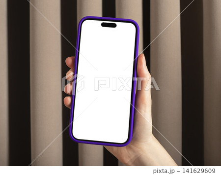 Hand holding smartphone with blank screen and purple case. Vertical mobile phone mockup against curtain background. Copy space for app advertising and product presentation.. Hand holding smartphone with blank screen and purple case. Vertical mobile phone mockup against curtain background. Copy space for app advertising and product presentation.. 141629609