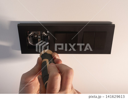 Hand holds a screwdriver to tighten a black electrical socket on a white wall. The person performs a 141629613