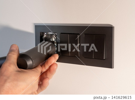 Hand installs a black electrical socket into a dark frame on a white wall. The electrician fits the Hand installs a black electrical socket into a dark frame on a white wall. The electrician fits the 141629615
