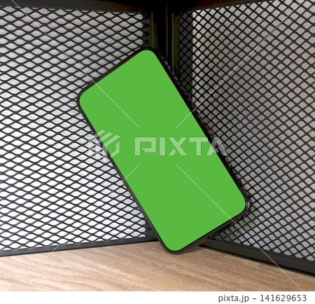 Phone mockup with a bright green chroma key screen leans diagonally in a wooden shelving corner. The Phone mockup with a bright green chroma key screen leans diagonally in a wooden shelving corner. The 141629653