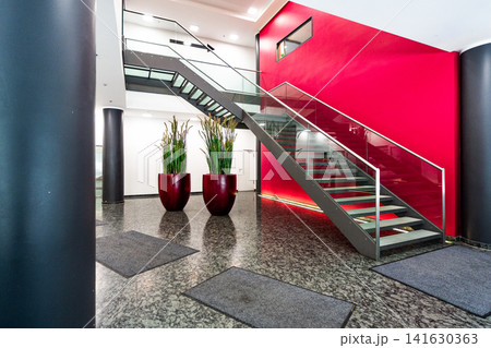 Modern office lobby with red feature wall and glass staircase, contemporary corporate interior with granite floor, cylindrical columns and large plant pots Modern office lobby with red feature wall and glass staircase, contemporary corporate interior with granite floor, cylindrical columns and large plant pots 141630363