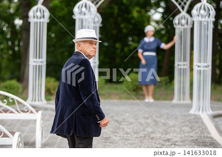 Elderly man in a hat walks towards a garden gazebo while a woman stands in the background on a sunny day Elderly man in a hat walks towards a garden gazebo while a woman stands in the background on a sunny day 141633018