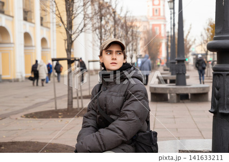 Young man in jacket and cap sitting on city street. Teenager feeling thoughtful in urban environment during autumn day. Lifestyle portrait of student waiting in outdoors public space. Young man in jacket and cap sitting on city street. Teenager feeling thoughtful in urban environment during autumn day. Lifestyle portrait of student waiting in outdoors public space. 141633211