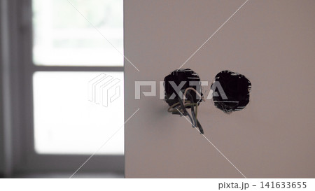 Exposed electrical outlet openings with loose wires on plain interior wall beside bright window light, showing unfinished renovation work, muted tones and empty room detail Exposed electrical outlet openings with loose wires on plain interior wall beside bright window light, showing unfinished renovation work, muted tones and empty room detail 141633655