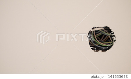 Damaged drywall surface with circular opening exposing tangled electrical wires inside wall cavity, unfinished interior detail with neutral beige background, construction damage Damaged drywall surface with circular opening exposing tangled electrical wires inside wall cavity, unfinished interior detail with neutral beige background, construction damage 141633658