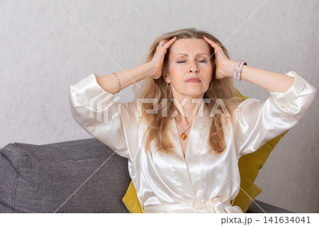 Middle-aged woman relaxing on a couch with eyes closed and hands on her head while wearing a silky robe in a neutral-toned room Middle-aged woman relaxing on a couch with eyes closed and hands on her head while wearing a silky robe in a neutral-toned room 141634041