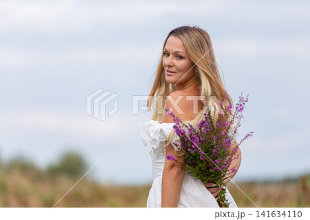 Woman in white dress holds a bouquet of wildflowers while looking back in a field during a cloudy day 141634110