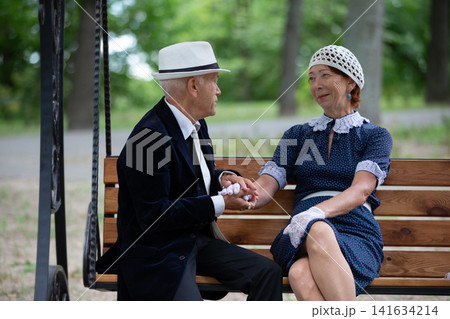 Elderly couple sitting on a bench in a park, enjoying a conversation and holding hands in the afternoon sun 141634214