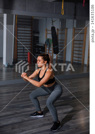 Young woman exercising with squats during focused gym fitness routine indoors. Beauty woman training lower body muscles with balanced athletic posture indoors. Strength wellness concept. Young woman exercising with squats during focused gym fitness routine indoors. Beauty woman training lower body muscles with balanced athletic posture indoors. Strength wellness concept. 141635186