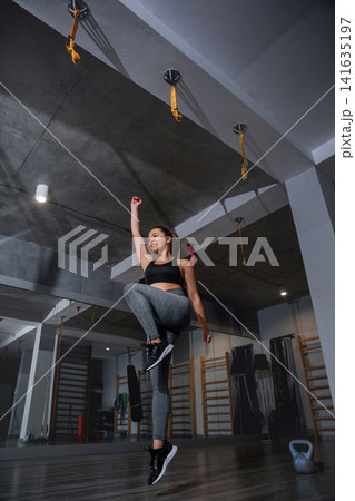 Beauty woman performing jumping knee raises during energetic gym routine indoors. Attractive woman training cardio endurance with healthy athletic posture inside studio. Fitness wellness concept. 141635197