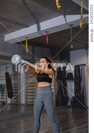 Young woman lifting kettlebell during energetic indoor fitness training routine. Pretty woman exercising upper body muscles with athletic balanced movement inside gym. Strength wellness concept. Young woman lifting kettlebell during energetic indoor fitness training routine. Pretty woman exercising upper body muscles with athletic balanced movement inside gym. Strength wellness concept. 141635203