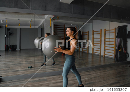Young woman holding fitness ball during energetic gym workout indoors. Pretty woman training core muscles with balanced athletic posture inside studio. Fitness wellness concept. Young woman holding fitness ball during energetic gym workout indoors. Pretty woman training core muscles with balanced athletic posture inside studio. Fitness wellness concept. 141635207