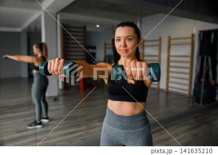 Beautiful woman holding dumbbells during active indoor fitness workout training. Young woman exercising arm muscles with healthy athletic posture inside gym. Strength wellness concept. Beautiful woman holding dumbbells during active indoor fitness workout training. Young woman exercising arm muscles with healthy athletic posture inside gym. Strength wellness concept. 141635210