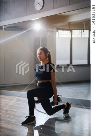 Attractive woman doing lunges inside bright modern fitness studio. Beauty woman exercising with dumbbells during active lower body training. Wellness gym concept. Attractive woman doing lunges inside bright modern fitness studio. Beauty woman exercising with dumbbells during active lower body training. Wellness gym concept. 141635261