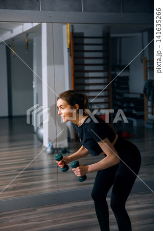 Beauty woman training with dumbbells in modern workout studio. Pretty woman performing athletic fitness exercises with healthy active posture. Wellness strength concept. 141635266