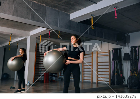 Beautiful woman holding fitness ball inside modern workout gym studio. Young woman preparing active exercise routine with healthy athletic posture indoors. Wellness sport concept. 141635278