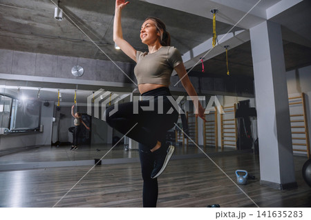 Woman practicing fitness moves in gym setting with exercise equipment in view and mirrors reflecting the activity during daytime Woman practicing fitness moves in gym setting with exercise equipment in view and mirrors reflecting the activity during daytime 141635283