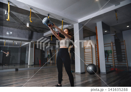 Woman exercises with a kettlebell in a modern gym during morning hours focused on strength training and fitness routine with gym equipment around her Woman exercises with a kettlebell in a modern gym during morning hours focused on strength training and fitness routine with gym equipment around her 141635288