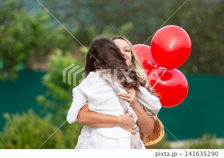 Emotional embrace between a mother and daughter with red balloons during a sunny outdoor event in spring Emotional embrace between a mother and daughter with red balloons during a sunny outdoor event in spring 141635290