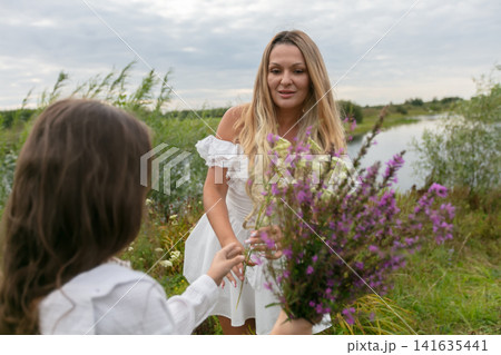 Woman in white dress receives flowers from child near serene river and greenery during overcast day 141635441