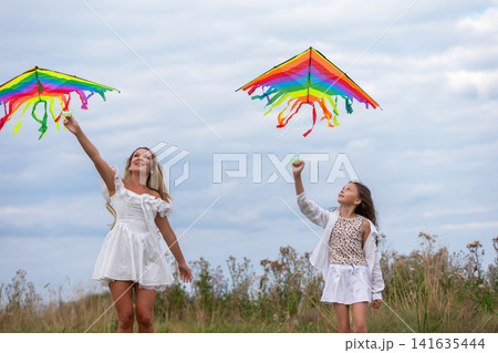 Two girls flying colorful kites in an open field on a cloudy day with soft grasses surrounding them 141635444