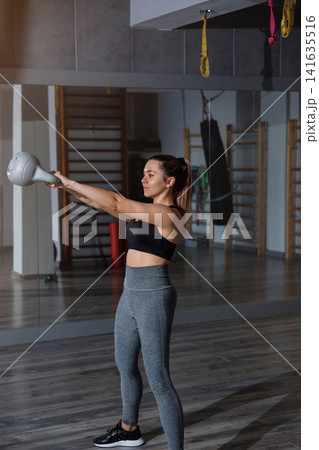 Young woman swinging kettlebell during active gym conditioning workout indoors. Pretty woman training upper body muscles with healthy athletic movement inside studio. Strength wellness concept. Young woman swinging kettlebell during active gym conditioning workout indoors. Pretty woman training upper body muscles with healthy athletic movement inside studio. Strength wellness concept. 141635516