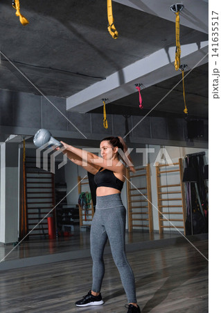 Attractive woman lifting kettlebell during indoor athletic fitness training session. Beauty woman exercising full body strength with balanced toned posture indoors. Wellness sport concept. Attractive woman lifting kettlebell during indoor athletic fitness training session. Beauty woman exercising full body strength with balanced toned posture indoors. Wellness sport concept. 141635517
