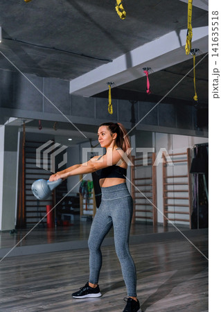 Pretty woman exercising with kettlebell during energetic indoor strength workout session. Young woman training arm muscles with athletic balanced movement inside gym. Fitness wellness concept. 141635518